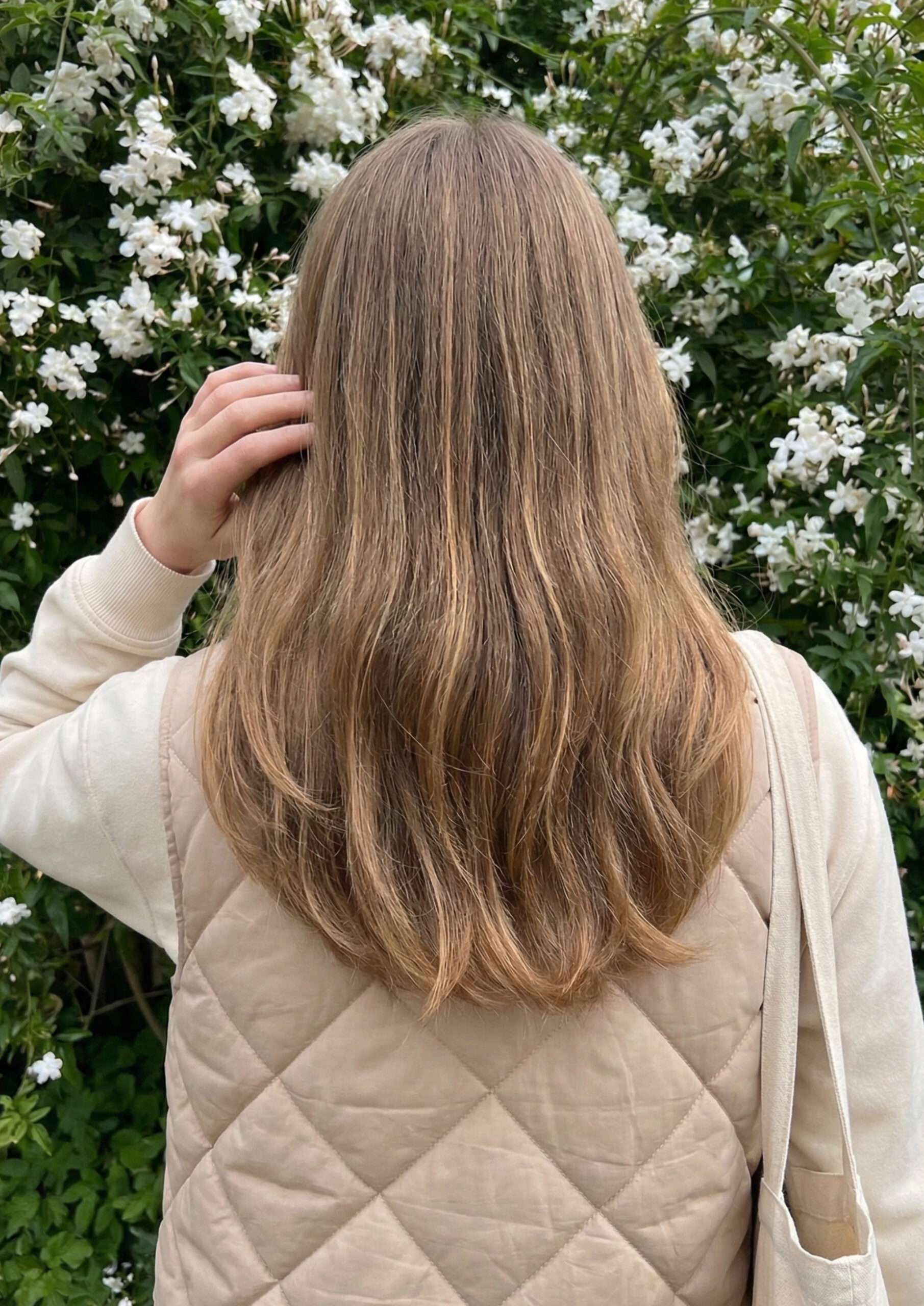 Mulher com cabelo loiro escuro acinzentado longo em U, em frente a um jardim